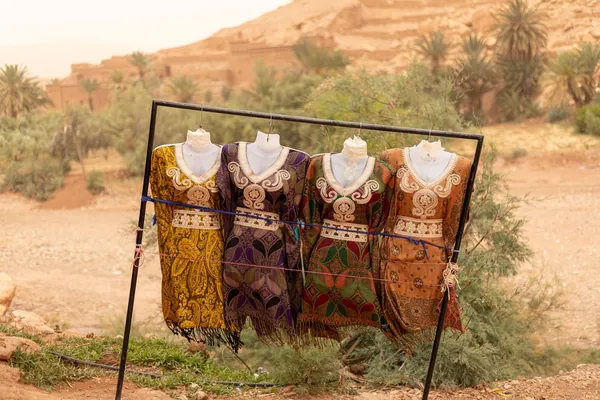 Quatre caftans brodés ocre, rouge et violet sur des bustes de mannequin, en plein air devant des palmiers
