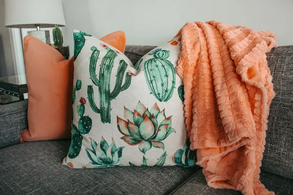 Coussin imprimé de cactus, coussin orange et plaid à franges posés sur un canapé gris