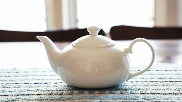 Théière en porcelaine blanche sur un set de table tissé bleu et blanc, devant une fenêtre lumineuse