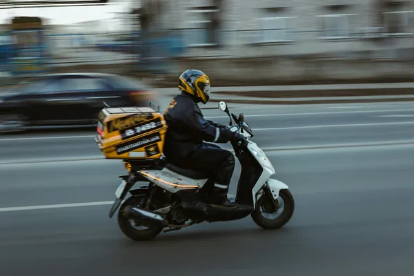 Livreur casqué sur un scooter avec un coffre de livraison jaune, dans une rue