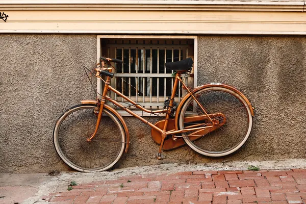 Vélo de ville ancien couleur cuivre appuyé contre un mur crépi, sous une fenêtre à barreaux