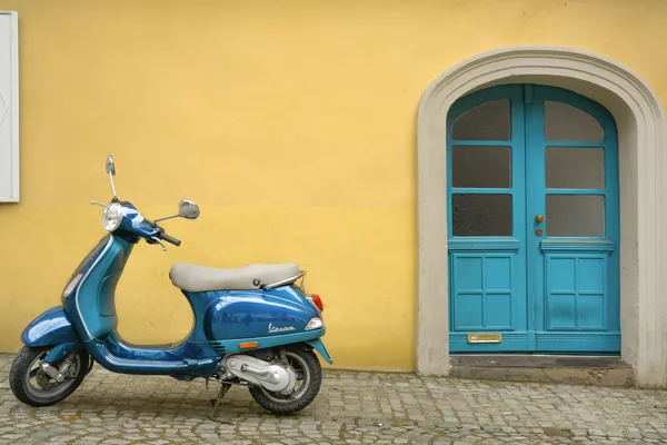 Scooter bleu de style rétro garé sur des pavés devant une façade jaune et une porte bleue
